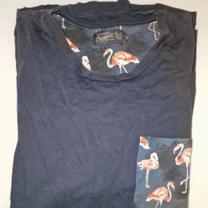 Mörkblå t-shirt med flamingomönster från Jack & Jones - Snygg mörkblå t-shirt med ett coolt flamingomönster på bröstfickan och insidan av ärmarna. Perfekt för en avslappnad stil med en touch av färg och mönster. Garderobsrensning så kika gärna på övriga auktioner.