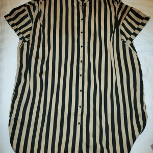 Randig kortärmad skjorta - Snygg kortärmad skjorta med vertikala ränder i svart och beige. Skjortan har knappar hela vägen ner och en avslappnad passform. Perfekt för en stilren look.