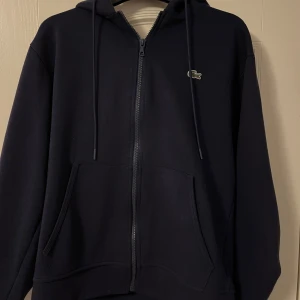 Mörkblå tröja från Lacoste - Säljer en stilren svart hoodie från Lacoste med dragkedja och huva. Tröjan har långa ärmar och en liten krokodil-logga på bröstet. Perfekt för en avslappnad look.