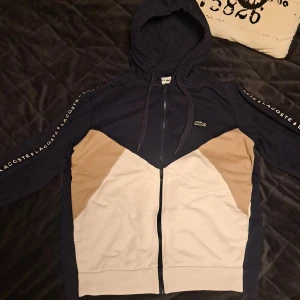 Blå och beige hoodie från Lacoste - Säljer en snygg blå och beige hoodie från Lacoste med dragkedja och huva. Tröjan har vita och beige paneler framtill och Lacoste-logga på ärmarna. Perfekt för en avslappnad stil. Hör av dig i pm vid funderingar✌️