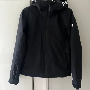 Svart skidjacka från Peak Performance - Snygg svart jacka från Peak Performance med huva och dragkedja. Jackan har en stilren design med logotyp på huvan. Knappt använd i storlek S.