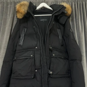  Cedrico - Säljer en svart dunjacka från Cedrico med pälskantad huva. Jackan har flera praktiska fickor med knappar och dragkedja framtill. Perfekt för kalla dagar med sin varma och skyddande design. 🖤storlek L 