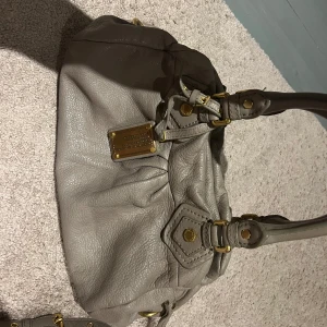 Grå handväska från Marc Jacobs classic q baby groove - Snygg grå handväska från Marc Jacobs i skinn med guldfärgade detaljer. Väskan har en stilren design med två handtag och en dekorativ platta framtill. Perfekt för att ge din outfit en lyxig touch.