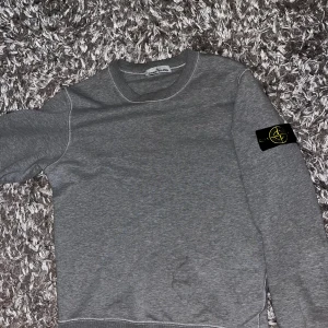 Grå tröja från Stone Island - Säljer en stilren grå tröja från Stone Island med deras ikoniska märke på ärmen. Tröjan har en klassisk rund hals och långa ärmar, perfekt för en avslappnad look. Passar bra till både jeans och chinos.