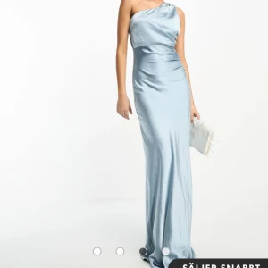 Ljusblå one-shoulder balklänning - Elegant ljusblå klänning med one-shoulder design. Klänningen har en lång och smickrande passform enda ner till fötterna, perfekt för speciella tillfällen. Materialet ger en fin glans och faller vackert längs kroppen. 