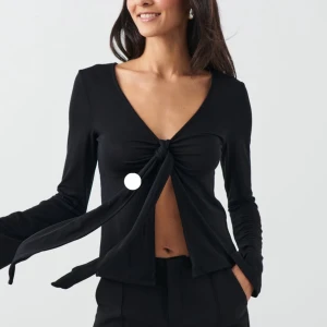 Svart långärmad knytblus - Snygg svart långärmad blus med knytning framtill. Perfekt för en stilren look med djup v-ringning och mjukt material. Från Gina liknade design by si säljer den på grund av att den inte kommer till användning längre 