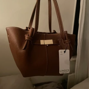 Brun handväska från Lindex - Snygg brun handväska från Lindex i syntetmaterial. Väskan har en elegant rosettdetalj och stängs med en knapp. Helt ny!🩷