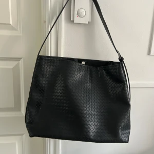 Svart axelväska med flätat mönster - Snygg svart axelväska med ett flätat mönster. Väskan har en justerbar axelrem med ett spänne i silver. Får plats med dator osv. Näst intill oanvänd 