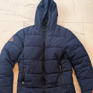 Syperdry jacka  - Säljer en blå pufferjacka från Superdry med huva och dragkedja. Jackan har ett broderat märke på ärmen och är perfekt för kyligare dagar. Den quiltade designen ger extra värme och stil. Köpt på pondus för 2-3 år sedan. Tvättas innan den skickas