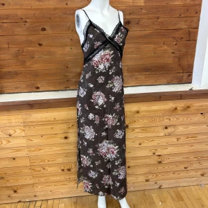 Blommig långklänning med spetsdetaljer - Säljer en elegant långklänning med blommigt mönster i rosa och gröna toner på en mörk bas. Klänningen har smala axelband och v-ringning med svarta spetsdetaljer. Perfekt för en somrig look!