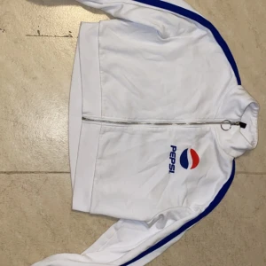 Vit jacka med Pepsi-logga - Jackan har blå ränder längs ärmarna och en hög krage. Perfekt för en sportig look!