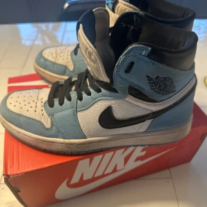 Nike Air Jordan 1 Retro High OG 'University Blue‘ - Säljer ett par Nike Air Jordans i en snygg kombination av blått och vitt. Skorna har en klassisk hög design med svart swoosh och snörning. Perfekta för dig som vill ha en stilren och sportig look.
