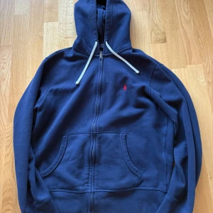 Ralph Lauren zip hoodie  - Säljer en snygg mörkblå hoodie från Polo Ralph Lauren med dragkedja och huva. Den har en liten röd logga på bröstet och praktiska fickor framtill. Perfekt för en avslappnad stil. Den har inga defekter alls, inga fläckar inga hål. Hör av dig vid fler frågor 