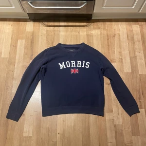 Mörkblå sweatshirt från Morris - Säljer en snygg mörkblå sweatshirt från Morris i storlek medium. Tröjan har ett tryck med texten 'Morris' och en brittisk flagga på bröstet. Perfekt för en casual look och passar bra till jeans. Tröjan är i bra skick och väldigt bekväm att ha på sig.