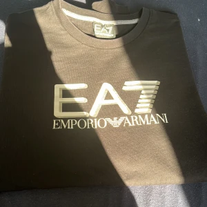 Svart tröja från Emporio Armani - Säljer en snygg svart tröja från Emporio Armani med deras ikoniska EA7-logga i vitt på bröstet. Tröjan är långärmad och perfekt för en stilren look. Passar både till vardags och fest!