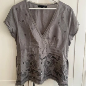 Grå blus med paljettdetaljer - Säljer en elegant grå blus från Vila med v-ringning och korta ärmar. Blusen har vackra paljettdetaljer och ett knytband i midjan för en fin passform. Perfekt för både vardag och fest!