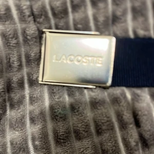 Blått bälte från Lacoste - Säljer ett snyggt blått bälte från Lacoste med en stilren silverfärgad spänne. Bältet har den klassiska Lacoste-krokodilen broderad i grönt och vitt. Perfekt för att ge en sportig touch till din outfit!