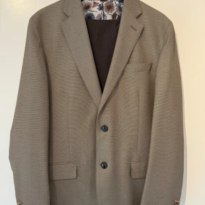 Beige kavaj från Dressmann - Säljer en snygg beige kavaj från Dressmann i storlek 52 (short). Kavajen har ett diskret mönster och är utrustad med två knappar framtill. Insidan är fodrad med ett blommigt mönster i blått och brunt. Perfekt för både fest och vardag!