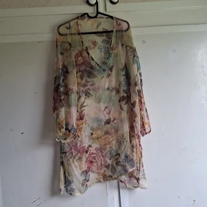 Blommig tunika med v-ringning - Säljer en superfin blommig tunika med v-ringning. Den är i ett tunt och luftigt material, perfekt för varma sommardagar. Tunikan har långa ärmar och ett löst passande snitt som gör den bekväm att bära. Färgerna går i ljusa toner med inslag av rosa, blått och grönt. Perfekt för en sommarfest eller en dag på stranden!