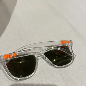 Transparenta solglasögon med orange text - Snygga solglasögon med transparent båge och mörka linser. På bågen står det sry ' i orange text. Perfekta för en solig dag och ger en cool look!