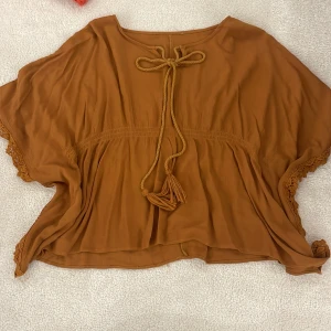 Brun topp med knytband och spetsdetaljer - Säljer en superfin brun topp med knytband framtill och vackra spetsdetaljer längs ärmarna. Toppen har en lös passform och är perfekt för en avslappnad stil. Materialet känns lätt och luftigt, vilket gör den idealisk för varmare dagar.