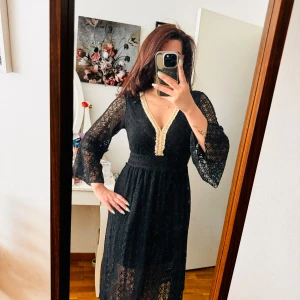 Svart spetsklänning med gulddetaljer - Säljer en superfin svart spetsklänning med gulddetaljer. Klänningen har en v-ringad hals med en vacker guldkant och långa ärmar. Den är perfekt för fest och har en tight passning i midjan som ger en smickrande siluett. Klänningen är i nyskick och är en långklänning som passar perfekt till höst och vinter.