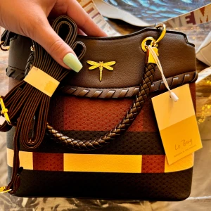 Randig handväska från Le Bag - Säljer en superfin handväska från Le Bag. Den är randig i brunt, gult och beige med en söt guldfärgad trollslända som detalj. Väskan är gjord av syntetmaterial och har en flätad axelrem. Perfekt för både vardag och fest!