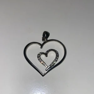 Hjärtaformad hängsmycke i silver - Säljer ett superfint hängsmycke i form av två hjärtan, ett större och ett mindre inuti, båda i silver. Det mindre hjärtat är dekorerat med glittrande stenar som ger en extra touch av elegans. Perfekt för att ge lite kärlek till din outfit! 💖