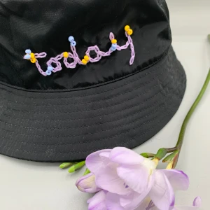 Svart hatt från & Other Stories - Säljer en superfin svart regnmössa från & Other Stories. Den har en cool handbroderad text 'idag' i lila med små gula och lila pärlor som detaljer. Perfekt för att ge din outfit lite extra stil! Storlek: M/56