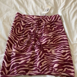 Zebramönstrad kjol med knytband - Säljer en supercool zebramönstrad kjol i lila och rosa. Den har ett knytband i midjan för perfekt passform och är gjord i ett mjukt och bekvämt material. Perfekt för en avslappnad dag eller en rolig fest!