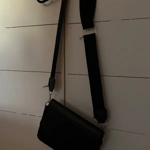 Svart axelväska med justerbar rem - Säljer en stilren svart axel eller crossover-väska i syntetmaterial. Väskan har en justerbar rem och stängs med magnetknappar. Perfekt för vardagsbruk eller en kväll ute. Den är rymlig nog för dina viktigaste saker och har en snygg, enkel design.