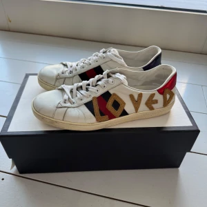 Gucci Ace ”Loved” - Storlek 42. Snygga vita sneakers från Gucci med texten 'LOVED' i guld på sidan. Skorna har röda och blå detaljer samt klassisk snörning. Inköpta i New York, kvitto finns med mitt namn.