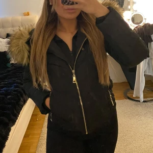 Svart parkajacka med pälshuva - Säljer en svart parkajacka med dragkedja och huva med pälsdetaljer. Jackan har långa ärmar och är perfekt för kyligare väder. Den har en stilren design med praktiska fickor.