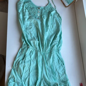 Turkos dress med spetsdetaljer - Säljer en söt turkos klänning med spetsdetaljer vid halsringningen och knappar framtill. Klänningen har en elastisk midja och är ärmlös, perfekt för varma dagar. Den har en avslappnad passform och är gjord i ett lätt material.