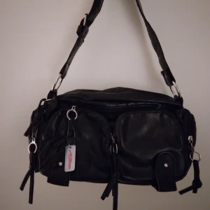 HELT NY & OANVÄND Svart praktisk handväska / väska med många fack. ARDENE - HELT NY & OANVÄND Svart praktisk handväska/axelväska med många fack, från kanadensiska märket ARDENE. Mjukt immitationsläder. Snygga silvriga detaljer. Inga defekter. Lapp/tag sitter kvar. Mått: Ca 31 x 20 x 10 cm. (Längd, höjd, djup)