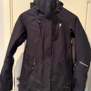 Svart jacka från Peak Performance - Säljer en svart vadderad  jacka från Peak Performance med dragkedja och huva.  Jackan har långa ärmar och flera praktiska fickor. Perfekt för skidåkning, utrustad med recco, putsduk för Googles och har en stilren design med logotypdetaljer. Vind och vatten tät, Hipe membran. Mycket fint skick,  använd endast några gånger pga fel storlek. Nypris 4500 kr. 