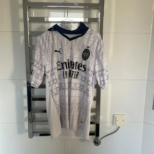 Vit AC Milan fotbollströja  - Säljer en vit AC Milan fotbollströja från Puma med grått mönster och svart krage. Tröjan har korta ärmar och klubbens emblem på bröstet. Perfekt för fans som vill visa sitt stöd. ⚽