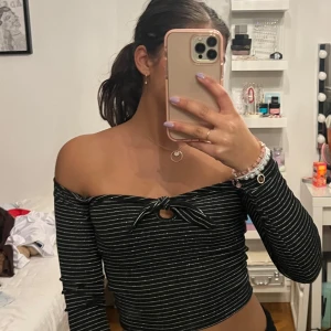 Svart offshoulder topp med knyt - Snygg svart glittrig offshoulder topp med vita ränder och knyt framtill. Toppen har långa ärmar och en liten öppning vid bysten. Perfekt för en stilren look.