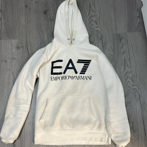 Vit hoodie från Emporio Armani - Säljer en stilren vit hoodie från Emporio Armani med det ikoniska EA7-logotypen tryckt på framsidan. Tröjan har en klassisk känguruficka och en justerbar huva med dragsko. Perfekt för en avslappnad och trendig look.