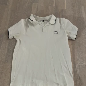 Vit pikétröja med svart kant - Snygg vit pikétröja med svart kant på krage och ärmslut. Tröjan har en liten broderad text på bröstet. Perfekt för en sportig look eller avslappnad stil.