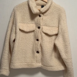  Teddyjacka    - Beige teddyjacka med knappar framtill. Jackan har en avslappnad passform och är perfekt för kyligare dagar. Den är långärmad och har en mjuk, fluffig yta som ger en trendig look.