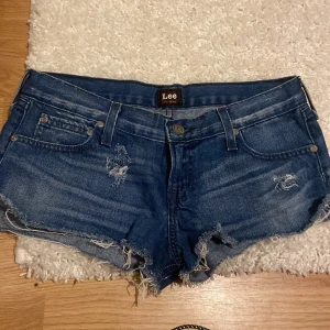Blå jeansshorts från Lee - Säljer ett par blå jeansshorts från Lee med slitna detaljer och fransar. Shortsen har en klassisk femficksdesign och knappgylf. Perfekta för en avslappnad stil.
