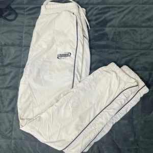 Vita träningsbyxor från FSBN - Säljer ett par vita träningsbyxor från FSBN med svart piping längs sidorna. Byxorna har elastisk midja med snörning och är perfekta för en sportig look. De är tillverkade i ett lätt och bekvämt material.