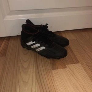 fotbollsskor Adidas - Säljer ett par svarta fotbollsskor från Adidas med vita ränder och snörning. Skorna har en högre ankel och är designade för bra grepp på planen. Perfekta för fotbollsträning och matcher.