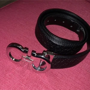 Svart bälte Ferregamo - Snyggt svart bälte från Salvatore Ferragamo med ett elegant mönster och en stilren silverfärgad spänne. Perfekt för att ge en lyxig touch till din outfit.