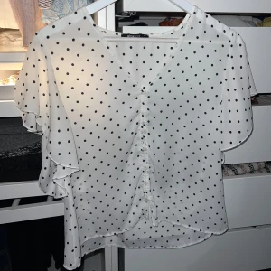 Vit prickig blus från Gina Tricot - Säljer en söt vit blus med svarta prickar från Gina Tricot. Blusen har en v-ringad design med volangdetaljer och knappar framtill. Skulle säga att den är något liten i storleken.