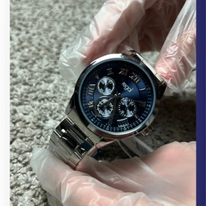 Silverfärgad klocka från Regal - (Priset går att sänka) Stilren klocka från Regal med en blå urtavla och romerska siffror. Klockan har en silverfärgad metallarmband och flera små urtavlor för extra funktioner. Perfekt för den som gillar en klassisk och elegant stil.