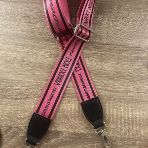 Rosa bälte från Don Donna - Snyggt rosa bälte från Don Donna med textdetaljer och justerbart spänne i silver. Perfekt för att ge en färgklick till din outfit. Bältet har svarta läderdetaljer vid ändarna och karbinhakar för enkel fastsättning.