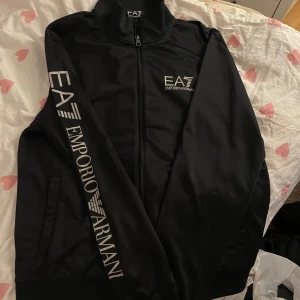 Svart jacka från Emporio Armani - Säljer en stilren svart jacka från Emporio Armani med EA7-logga på bröstet och ärmen. Jackan har en dragkedja framtill och är långärmad. Perfekt för en sportig look eller en avslappnad dag ute.