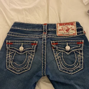 True Religion jeans - Snygga blå jeans från True Religion med skitsnygga röda detaljer och snygga visa sömmar. Byxorna har en extra låg midja jätte snygga fickor med knappar och broderade bakfickor. Sömmen på ena fickan är lite av men går jätte lätt att sy 💕 pris kan diskuteras 